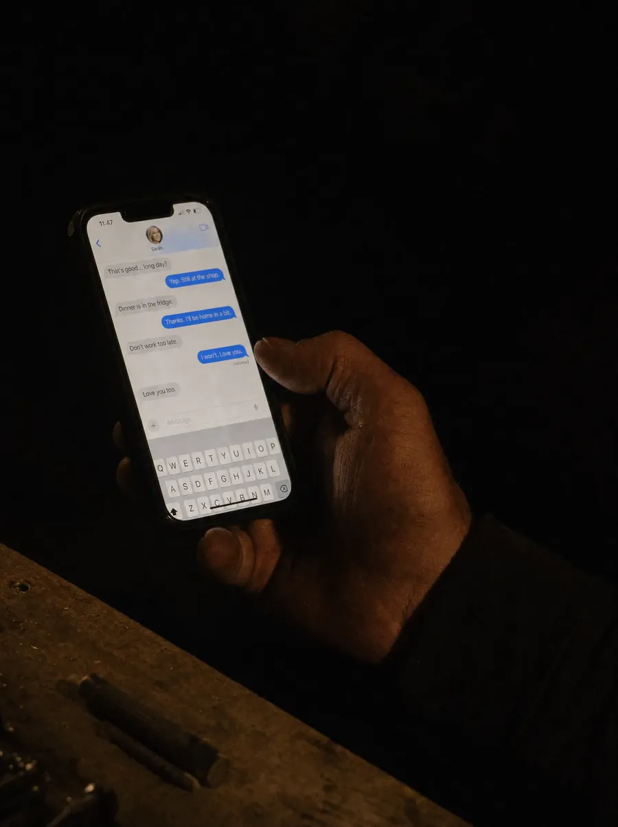 A hand holding a phone in a dark workshop after hours, a messaging thread still running.